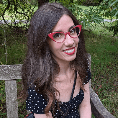 A Krissia é uma mulher branca e magra, de cabelo liso castanho e pontas onduladas. Ela usa um óculos vermelho, vestido preto de bolinhas e está sorrindo.